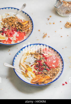 Healthy breakfast yogurt bowls with granola, grapefruit, banana, pomegranate, dried fruit, nuts and seeds over grey marble table background, selective Stock Photo