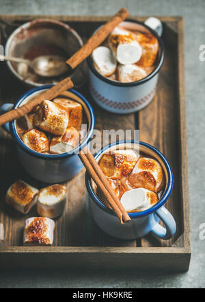 Hot chocolate in enamel mugs with cinnamon and roasted marshmallows in wooden tray over grey background, selective focus Stock Photo