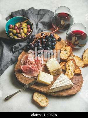 Wine and snack set. Variety of cheese, olives, prosciutto, roasted baguette slices, grapes on wooden board and glasses of red wine over grey marble ba Stock Photo