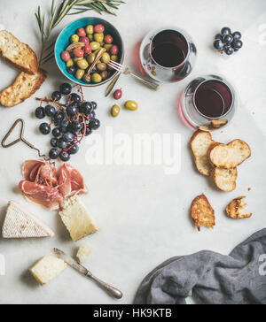 Wine and snack set. Variety of cheese, olives in ceramic bowl, prosciutto, roasted baguette slices, black grapes and glasses of red wine over grey mar Stock Photo