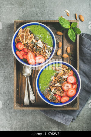 Healthy breakfast. Green smoothie bowls with strawberries, granola, chia and pumpkin seeds, dried fruit and nut in wooden tray over grey concrete back Stock Photo