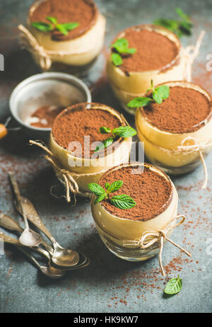 Homemade Italian dessert Tiramisu served in individual glasses with mint leaves and cocoa powder over grey concrete background, selective focus, verti Stock Photo