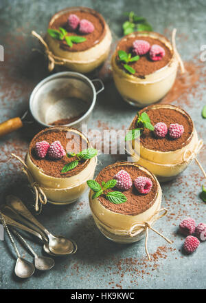 Homemade Italian dessert Tiramisu served in individual glasses with frozen raspberries, fresh mint leaves and cocoa powder over grey concrete backgrou