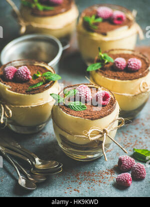 Homemade Italian dessert Tiramisu served in individual glasses with frozen raspberries, fresh mint leaves and cocoa powder over grey concrete backgrou