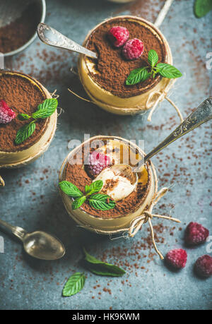 Homemade Italian dessert Tiramisu served in individual glasses with fresh mint leaves, raspberries and cocoa powder over grey concrete background, top Stock Photo