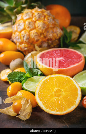 Variety of whole and sliced citrus fruits pineapple, grapefruit, lemon, lime, kumquat, clementine and physalis with mint over dark wooden background. Stock Photo