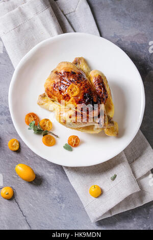 Whole roasted mini chicken cooking with caramelized kumquats served on white plate with textile napkin and fresh citrus fruits over gray kitchen table Stock Photo
