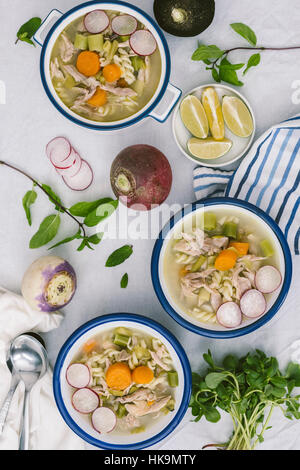 Three bowls of chicken soup with leek, carrot and pasta served with turnips, radishes, lemon wedges, mint and purslane. Stock Photo