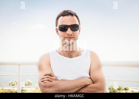 Young adult sporty Caucasian man in white shirt and black sunglasses stands with crossing hands. Outdoor summer portrait with sea on a background Stock Photo