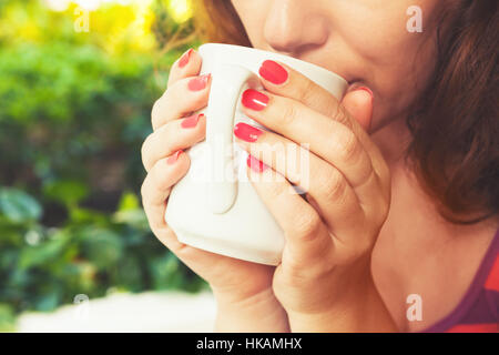Young Caucasian woman drinks coffee from big white cup. Close-up outdoor photo, selective focus on hands Stock Photo
