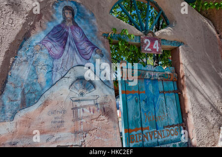 Religious mural of Jesus on a rustic adobe wall in Chimayó, New Mexico.  The turquoise gate reads Bienvenidos or Welcome. Stock Photo