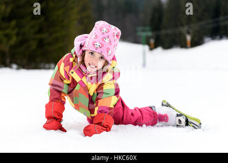 Little girl in winter outfit fell while skiing. Kid is lying in the snow with skis smiling. Happy ski experience in resort. Skiing accident. Stock Photo