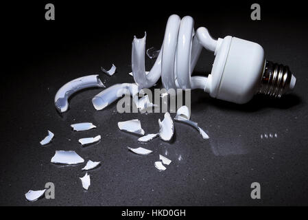 A Broken Compact Fluorescent bulb lays shattered in shards of glass on a hard surface with a dark background Stock Photo