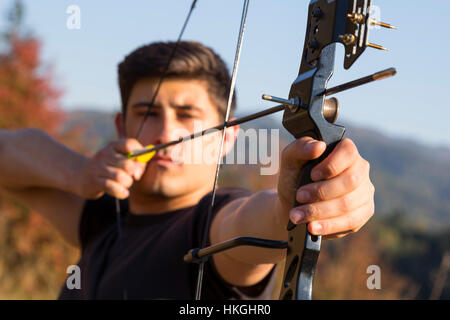 An archer draws his compound bow and aims upwards with clean blue sky as background. Stock Photo