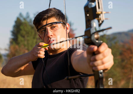 An archer draws his compound bow and aims upwards with clean blue sky as background. Stock Photo