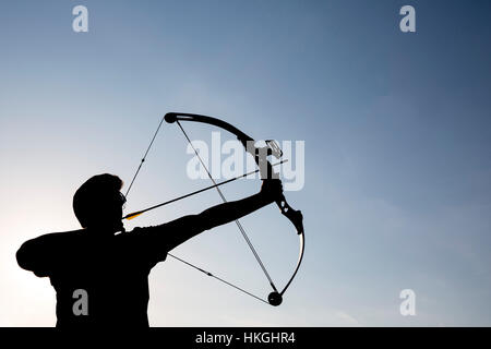 A silhouette of an archer draws his compound bow and aims upwards with clean blue sky as background. Stock Photo