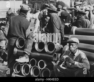 Black and white photograph of a group of African-American workers for an Emergency Defense Office, a United States federal emergency war agency set up to coordinate state and federal measures for protection of civilians in case of war emergency, housing construction job, during lunch break, standing around and sitting on large pipes, Washington, DC, December, 1941. From the New York Public Library. Stock Photo