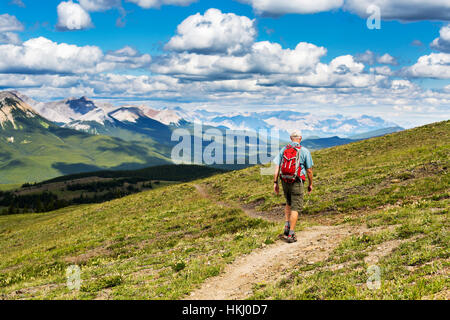 Male hiker walking along a hillside trail overlooking mountain range and valley with blue sky and cloud, West of Bragg Creek; Alberta, Canada Stock Photo