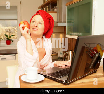 Young woman in bath robe using laptop and eating an apple Stock Photo