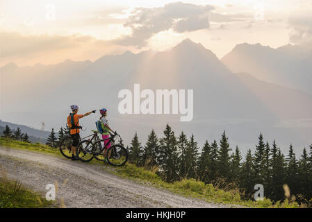 Young couple of mountain bikers standing on dirt road and looking at view during sunset, Zillertal, Tyrol, Austria Stock Photo