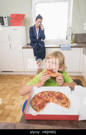 Boy eating pizza while mother has business call in kitchen,Bavaria,Germany Stock Photo