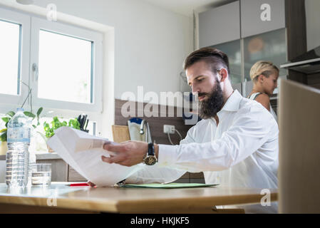 Young man with documents at the kitchen table and woman working in the background, Bavaria, Germany Stock Photo