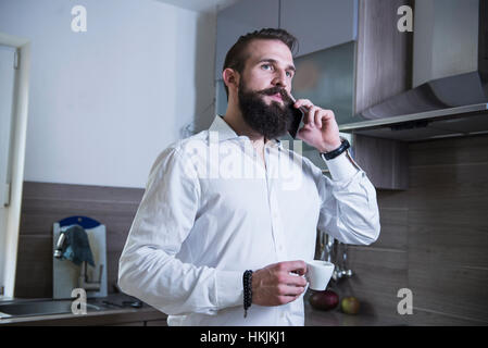 Young man talking on smartphone and drinking coffee in the kitchen, Bavaria, Germany Stock Photo