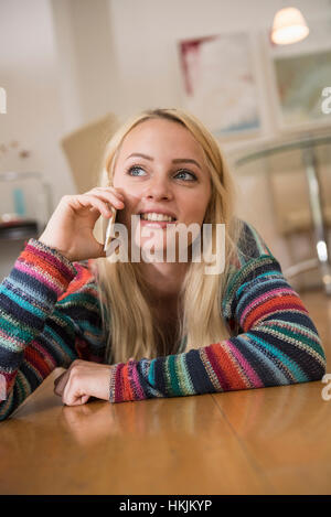 Beautiful young woman talking on smart phone and lying on floor in the living room, Bavaria, Germany Stock Photo
