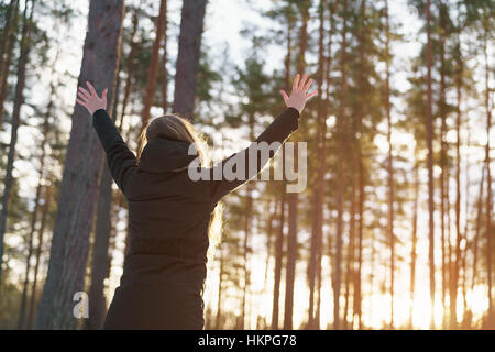 teen girl raised hands from behind in winter pine forest in sunset Stock Photo