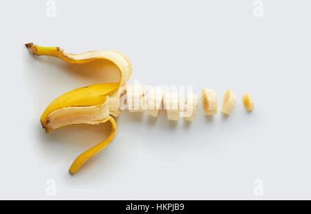 A semi peeled yellow banana cut into slices on a white surface Stock Photo