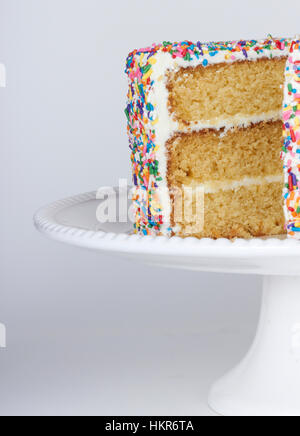 White cake on a cake stand covered in white frosting and rainbow nonpareil sprinkles with a slice missing Stock Photo
