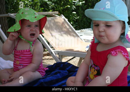 funny photo of baby girls wearing swimsuits and sun hats sitting in summer sun looking unhappy, funny kids concept children