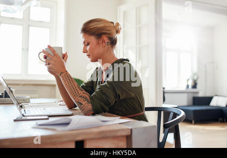 Side view shot of thoughtful young woman with coffee sitting at table. Female sitting at home office looking serious. Stock Photo