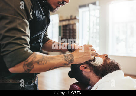 Professional barber holding brush and applying foam on the face of his client while going to shave. Hairdresser shaving customer in salon. Stock Photo