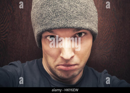 Young agressive Caucasian man in gray hat. Close-up studio face portrait over dark wooden wall background, selective focus Stock Photo