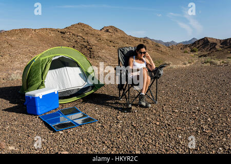 Woman in the desert calling while her smartphone is charging with a portable and foldable solar panel. Green energy concept Stock Photo