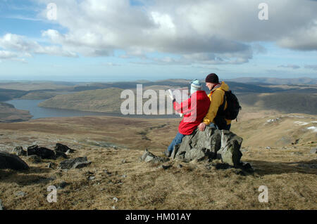 Two hill walkers sitting on rocks at summit reading map on bright winters day with lake in far distance Stock Photo