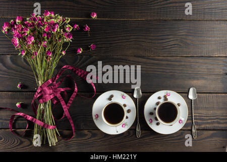Bouquet of dry flowers with a ribbon on the dark wooden surface. Two porcelain cups of coffee and two saucers Stock Photo