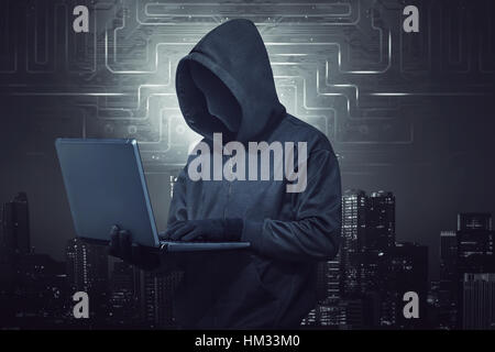 Hooded hacker holding laptop while typing against digital city background Stock Photo