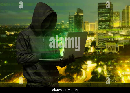 Hacker man with mask typing on laptop against modern city background Stock Photo