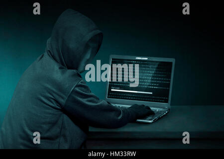 Hooded hacker with anonymous mask using laptop to steal data in the dark room Stock Photo