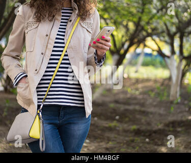 The girl standing on the street in the spring on the background of trees, dressed in a beige jacket, blue jeans and a striped shirt. In her left hand  Stock Photo