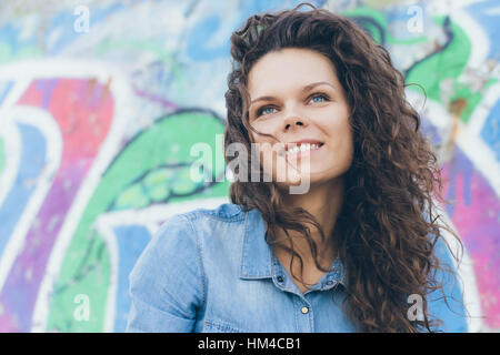 Portrait of happy smiling woman with blue eyes and curly hair in a denim shirt on a background of graffiti. Dreamy look up. Stock Photo