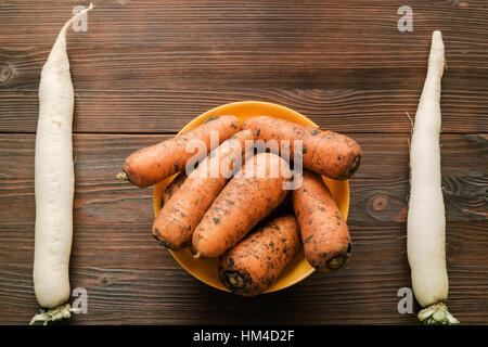 Unpeeled carrots in a bowl and daikon radish on a wooden table, top view Stock Photo