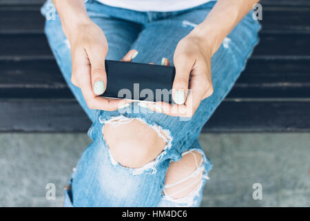 Girl in torn jeans sitting on a bench and uses a touch screen mobile phone. Top view close-up. Stock Photo