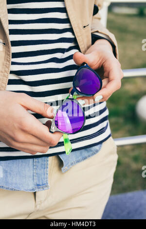 Woman in coat and striped t-shirt holding bright colored sunglasses Stock Photo