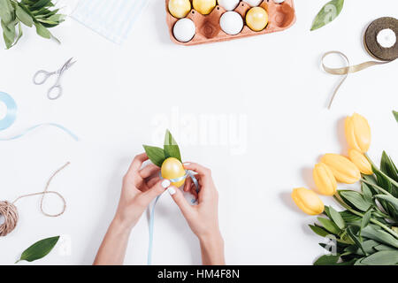 Female hand bandage yellow painted egg blue ribbon. On the table lay a tray with colored eggs, tulips, rope, ribbon and scissors, top view. Stock Photo
