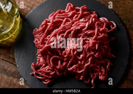 raw beef ground meat on a slate plate and a glass cruet with olive oil, on a rustic wooden table Stock Photo
