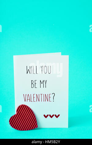 a red heart and a homemade postcard, made by myself, with the text will you be my valentine written in it, on a bright blue background Stock Photo