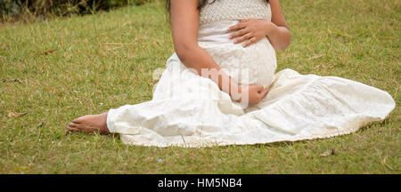 A pregnant woman is posing in the outdoor wearing a beautiful white dress. Stock Photo